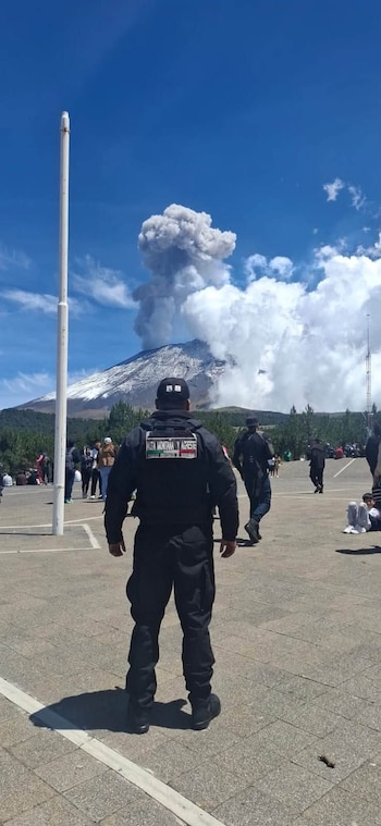 Hombre de espaldas con uniforme negro y gorra observa un volcán nevado con columna de humo. Cielo azul, nubes. Personas y árboles al fondo. Superficie pavimentada