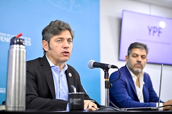 Primer plano de Axel Kicillof hablando ante un micrófono, con Carlos Bianco borroso detrás. Hay una pantalla con 'YPF' y un cartel azul