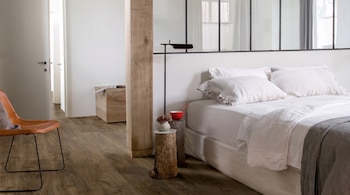 Vista de un dormitorio moderno con cama vestida de blanco, dos mesitas de noche de madera, una lámpara negra, una silla de cuero y suelo de madera oscura.