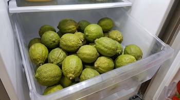 Un cajón transparente de un refrigerador lleno de limones verdes con la piel seca y arrugada, algunos con manchas oscuras, vistos desde arriba.