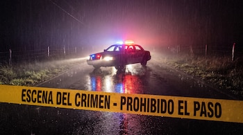 Cinta amarilla con el texto ESCENA DEL CRIMEN - PROHIBIDO EL PASO delante de un patrullero con sirenas encendidas en una vía mojada.