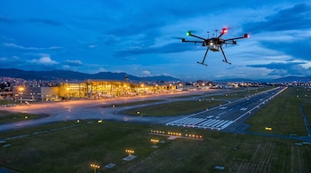 Dron con luces rojas y verdes flotando sobre una pista de aeropuerto iluminada al anochecer, con el edificio terminal y luces urbanas de Bogotá al fondo.