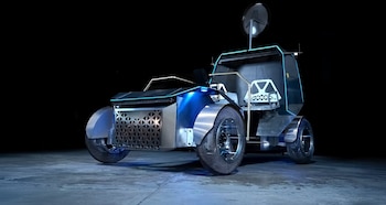 Un prototipo de vehículo lunar plateado y negro con luces azules y ruedas de malla oscura, estacionado sobre un suelo de hormigón gris