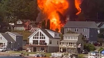 Una enorme columna de llamas y humo naranja y rojo se eleva sobre las casas, con un denso bosque verde en el fondo