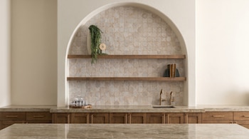 Interior de cocina moderna con nicho arqueado empotrado. Cuenta con azulejos geométricos claros, estantes de madera, grifos de bronce y encimeras de mármol beige.
