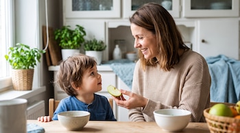 Adulto y niño pequeño sentados a una mesa de madera en una cocina, el adulto sostiene una rodaja de manzana verde, hay cuencos y plantas en macetas.