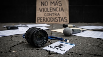Primer plano de un lente de cámara roto y una credencial de prensa sobre asfalto agrietado. Un micrófono, papeles y un cartel borroso se ven al fondo.