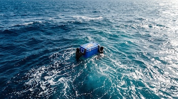 Vista aérea de dos personas en mar abierto sujetando los laterales de una hielera plástica azul.