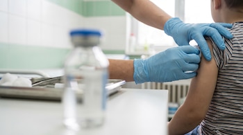 Dos manos enguantadas de celeste vacunan el brazo de un niño, con un vial de vacuna desenfocado en primer plano y un fondo hospitalario verde y blanco.