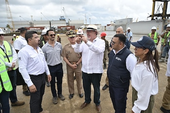 Grupo de hombres y mujeres en un puerto. Un hombre con sombrero y camisa blanca apunta con las manos. Contenedores de carga, grúas portuarias y chalecos de seguridad