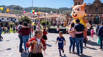 Plaza con personas, niños y adultos, un personaje inflable de animal, bailarines con vestimenta tradicional, globos, serpentinas y edificios coloniales.