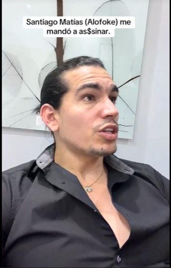 Hombre con cabello largo recogido y barba ligera, vestido con una camisa negra desabrochada y collar, hablando y mirando hacia la derecha