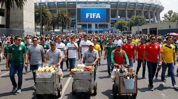 Vista general de una multitud de personas caminando hacia el Estadio Ciudad de México, con el logo de FIFA en una pantalla grande. Vendedores ambulantes en primer plano ofrecen comida y bebidas.