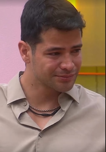Primer plano de un hombre con pelo oscuro corto, camisa beige y dos collares, uno plateado y otro negro. Sus ojos están humedecidos