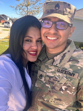 Primer plano de un hombre con uniforme militar del Ejército de EE. UU. y gafas, sonriendo junto a una mujer de cabello oscuro y blusa blanca, ambos sonriendo. Un vecindario se ve al fondo