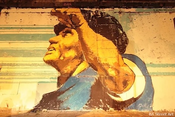 Mural de un hombre, Diego Maradona, con un brazo levantado. Tonalidades ocre, marrón, azul y blanco. Pared con franjas. Firma Lean Frizzer