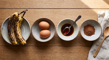Cuatro recipientes alineados en una mesa contienen dos plátanos, dos huevos, esencia de vainilla y canela en polvo.
