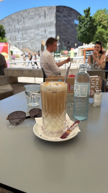 Mesa de café al aire libre con un café helado, vaso de agua, botella de agua, gafas de sol y un azucarero. Al fondo, un edificio moderno y gente borrosa