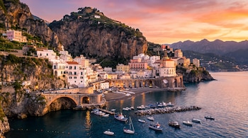 Pueblo costero con edificios blancos y de colores sobre una colina rocosa. Hay un puente, una playa, barcos en el mar y montañas al fondo bajo un cielo naranja.