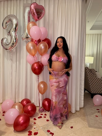 Lyss Okada embarazada con top y falda estampados posa frente a globos rosas, rojos y plateados con los números 2 y 1, y pétalos en el suelo