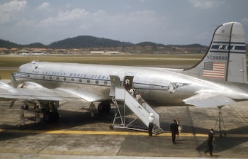Un avión plateado Pan American World Airways Clipper Seafarer con escaleras y pasajeros, personas en pista, edificios y colinas al fondo