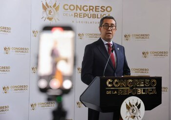 Hombre con traje y anteojos habla en un podio del Congreso de la República con un teléfono celular desenfocado en primer plano