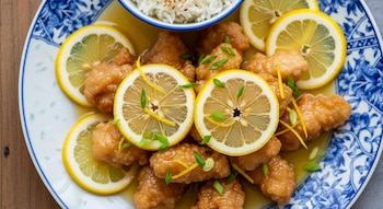 Pollo frito bañado en salsa de limón, con rodajas de limón, ralladura de limón y cebolla verde, servido con arroz blanco en un tazón, sobre un plato.
