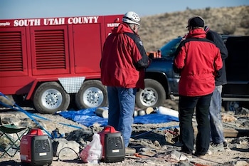 Tres personas de espalda, trailer rojo con texto 'SOUTH UTAH COUNTY', camioneta oscura, dos generadores portátiles rojos, lona azul, terreno árido