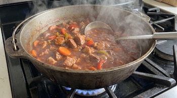 Olla de hierro sobre un quemador de gas encendido con estofado de carne y verduras emanando vapor, con un cucharón de metal dentro.