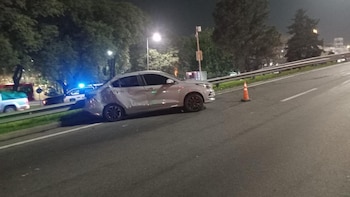 Automóvil sedán plateado con daños en carrocería, cono de tránsito naranja, vehículo policial con luces azules intermitentes, árboles, carretera