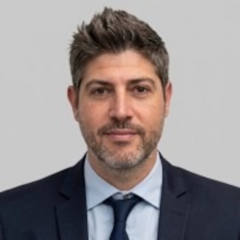 Retrato de un hombre con traje oscuro, camisa clara y corbata azul sobre un fondo gris claro