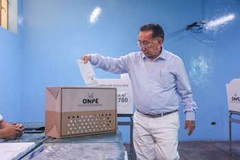 Hombre con camisa celeste y lentes depositando un voto en una urna de cartón con el logo de ONPE, en un centro de votación con paredes azules