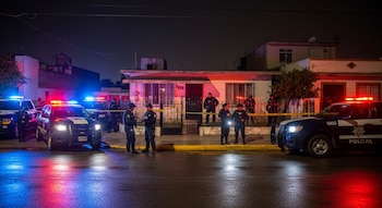 Varios vehículos policiales con luces encendidas y agentes de policía frente a una casa acordonada con cinta amarilla en una calle de noche.
