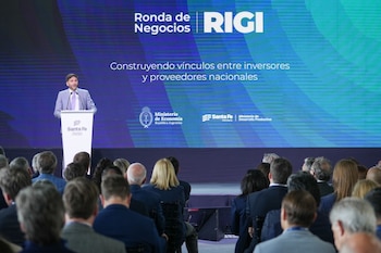 Un hombre de traje habla desde un atril ante una audiencia sentada de espaldas frente a una pantalla con texto institucional