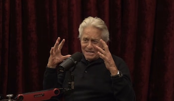 Michael Douglas gesticula con las manos abiertas frente a un micrófono en un estudio con cortinas de color rojo oscuro