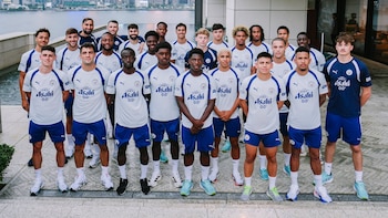 Grupo de veinte futbolistas hombres con uniformes blancos y azules del Manchester City. Posan al aire libre frente a un edificio y un cuerpo de agua