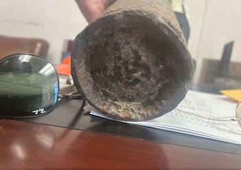 Objeto metálico cilíndrico oxidado sobre pila de papeles en un escritorio. A su izquierda hay unas gafas de sol negras. Se observa una mano en la parte superior