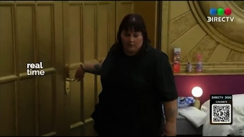 Una mujer de cabello oscuro y camiseta negra manipula una puerta dorada en una habitación. Al fondo, hay una pared púrpura, una lámpara y parte de una cama