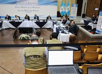 Vista de sala de conferencias con personas sentadas en mesas, observadores, banderas de Guatemala, y un hombre con chaleco de la OEA frente a una laptop