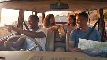Cuatro amigos sonrientes en un coche. El conductor señala, otro hombre mira un mapa, y dos mujeres en la parte trasera comparten aperitivos. La carretera está al fondo.