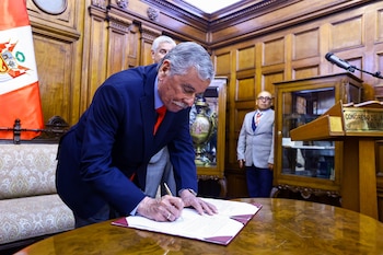 Hombre de cabello canoso en traje azul y corbata naranja firma un documento sobre una mesa de madera, con la bandera peruana y paneles de madera de fondo