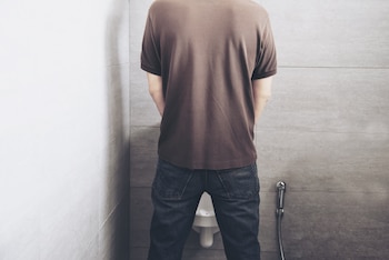Un hombre de espaldas en un urinario público con camiseta marrón y jeans azules. Las paredes son de azulejo gris y se ve una manguera de bidé