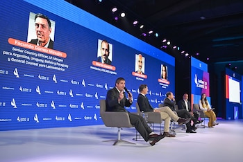 Cinco panelistas sentados en un escenario durante el AmCham Summit 26, con una gran pantalla LED azul de fondo mostrando los nombres de los oradores