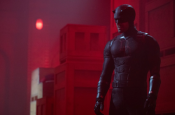 Un hombre musculoso con un traje negro ajustado y un casco con cuernos de diablo se para en un espacio oscuro iluminado por una fuerte luz roja