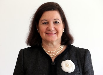 Mujer de cabello oscuro con collar de perlas y broche de flor blanca en chaqueta negra, sonríe a cámara sobre fondo blanco