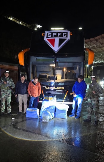 Un autobús negro con el logo SPFC, cuatro hombres y dos agentes uniformados de pie, junto a varios paquetes en el suelo mojado por la noche