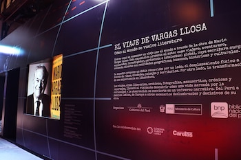 Panel de exposición oscuro con texto "EL VIAJE DE VARGAS LLOSA", un retrato sonriente en blanco y negro de Mario Vargas Llosa, y logos institucionales