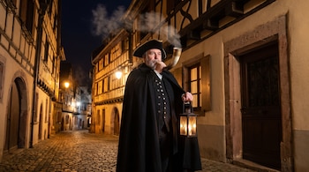 Un hombre con barba, capa negra, tricornio, farol encendido y cuerno de caza en una calle empedrada de noche entre casas tradicionales de madera.