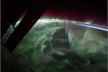 La curvatura de la Tierra iluminada por luces verdes y púrpuras, con un cielo estrellado oscuro y parte de un panel solar en primer plano