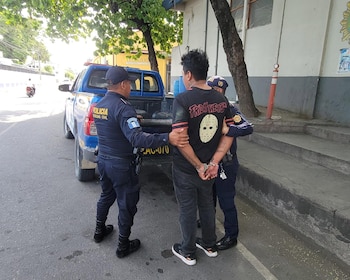 Dos policías con uniforme azul y un hombre esposado con camiseta negra. Detrás hay una camioneta azul en una calle arbolada y edificios al fondo